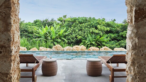 A pool area with two wooden lounge chairs and two woven stools, surrounded by a concrete deck and a view of lush greenery. - The Rescape (Miyakojima)