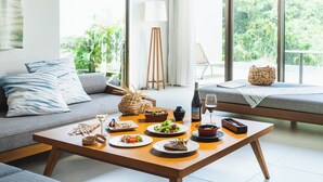 A modern living room with a dining table set for a meal, a sofa, and a large window with a view of greenery. - The Rescape (Miyakojima)
