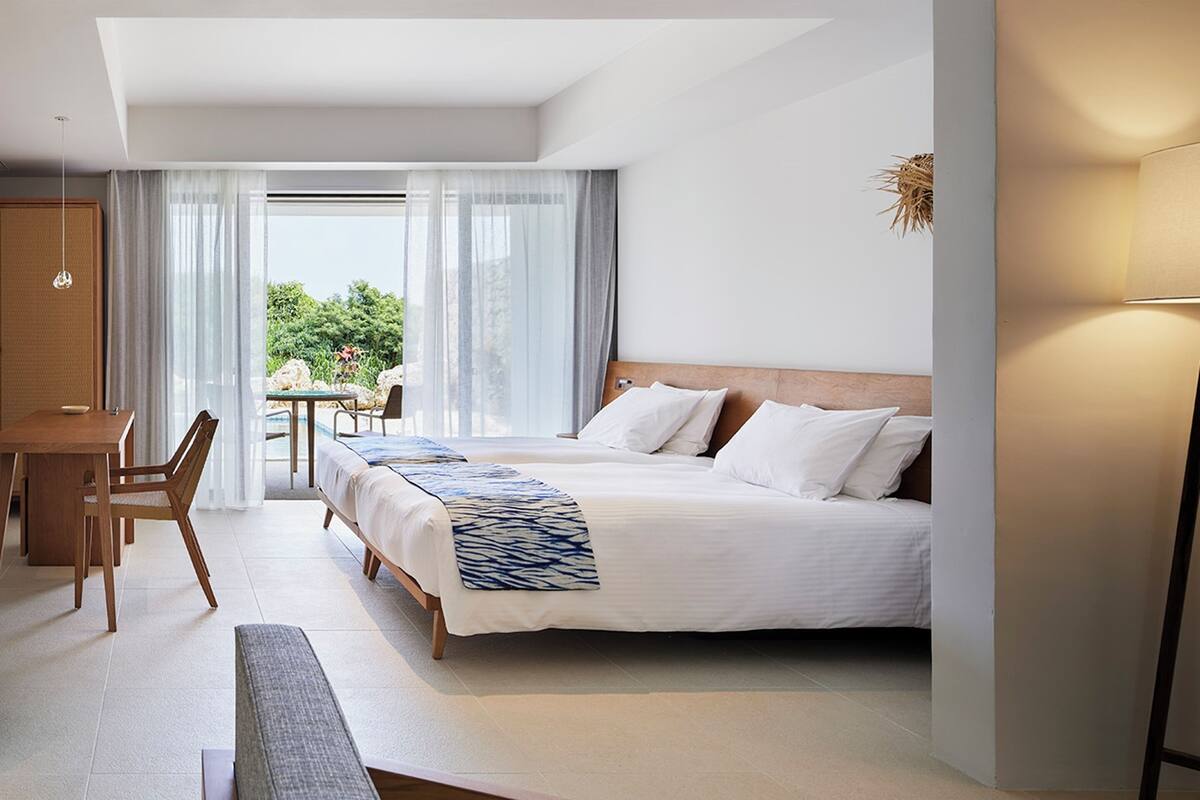 A hotel room with a bed, a desk, and a chair. There is a view of greenery outside the window.