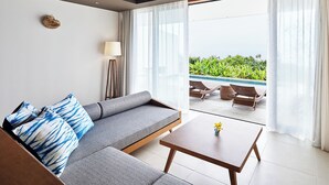 A modern living room with a grey sofa, a small wooden table, and a floor lamp. A large sliding glass door leads to an outdoor area with a pool and greenery. - The Rescape (Miyakojima)