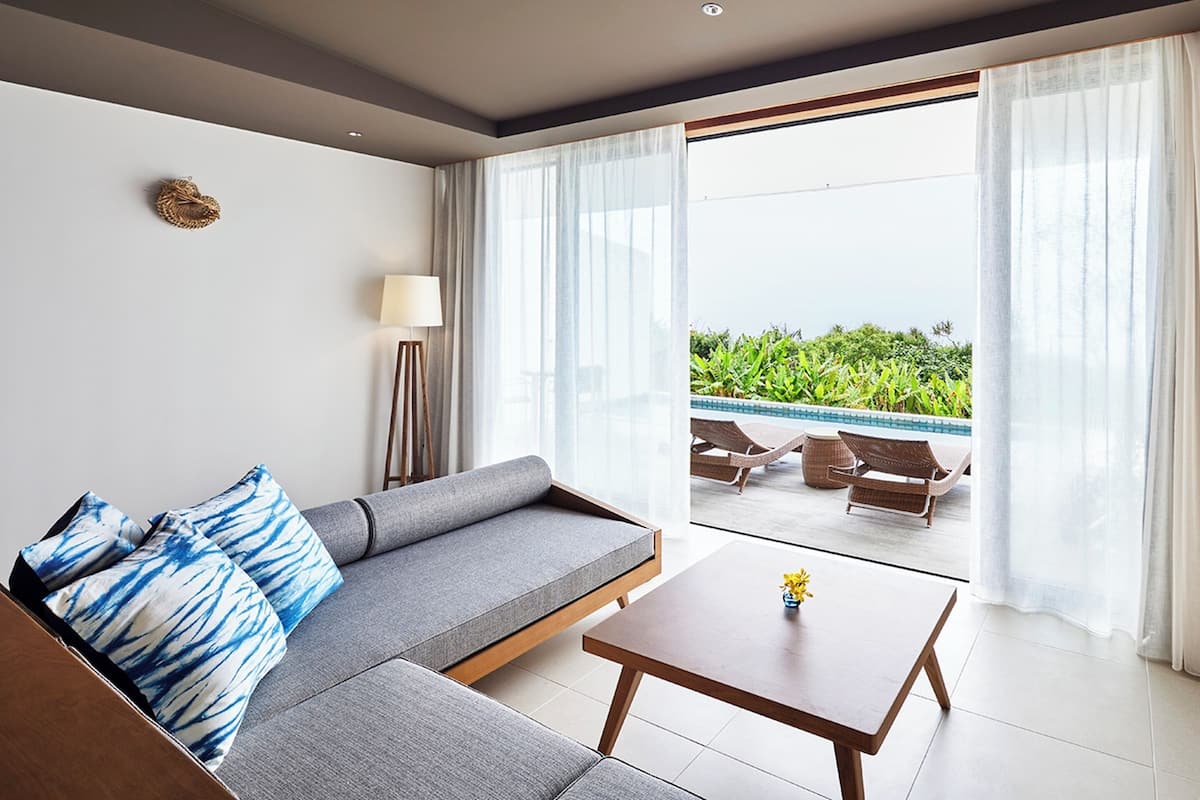 A modern living room with a grey sofa, a small wooden table, and a floor lamp. A large sliding glass door leads to an outdoor area with a pool and greenery.