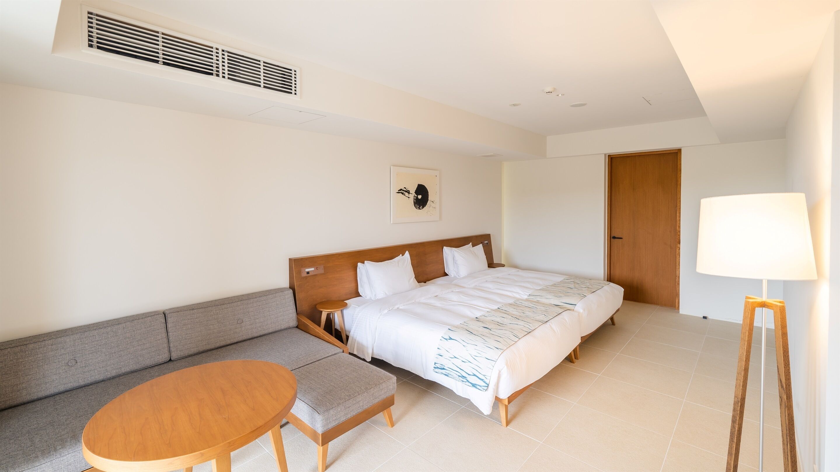 A hotel room with a bed, a sofa, a small table, a lamp, and a ceiling vent.
