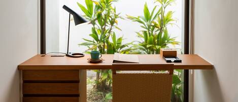 A wooden desk with a lamp, a cup, and a book, positioned by a window with plants.