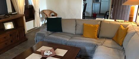 A living room with a sofa, a coffee table, a TV, and a kitchen visible through an archway.
