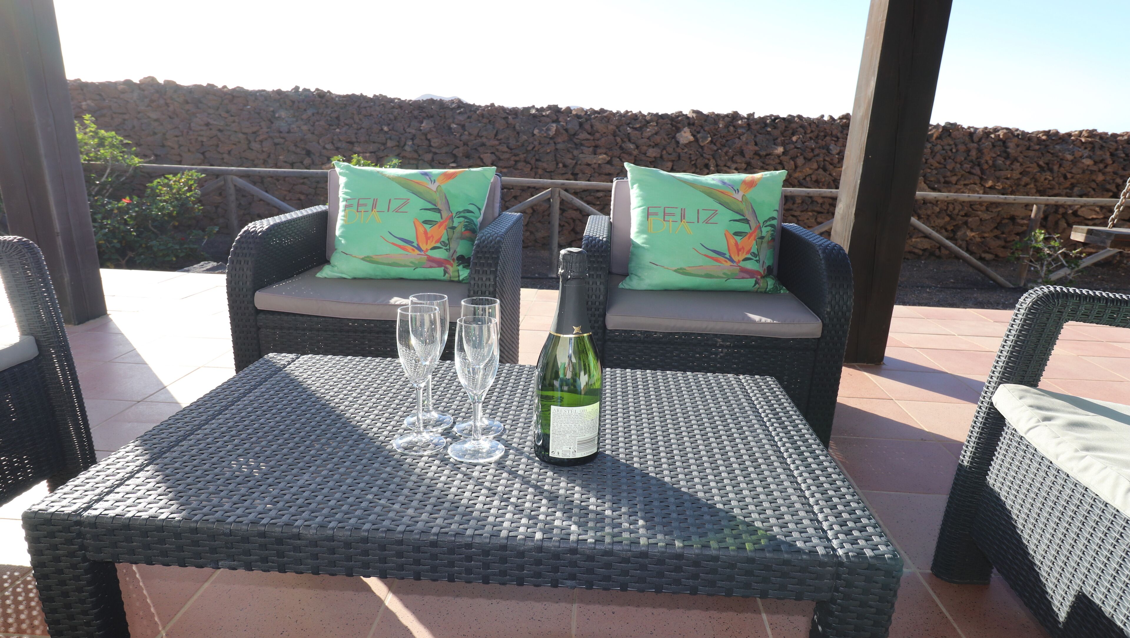 Une table en osier, avec deux verres à vin et une bouteille de vin, est placée sur une terrasse agrémentée de chaises et de coussins en osier.