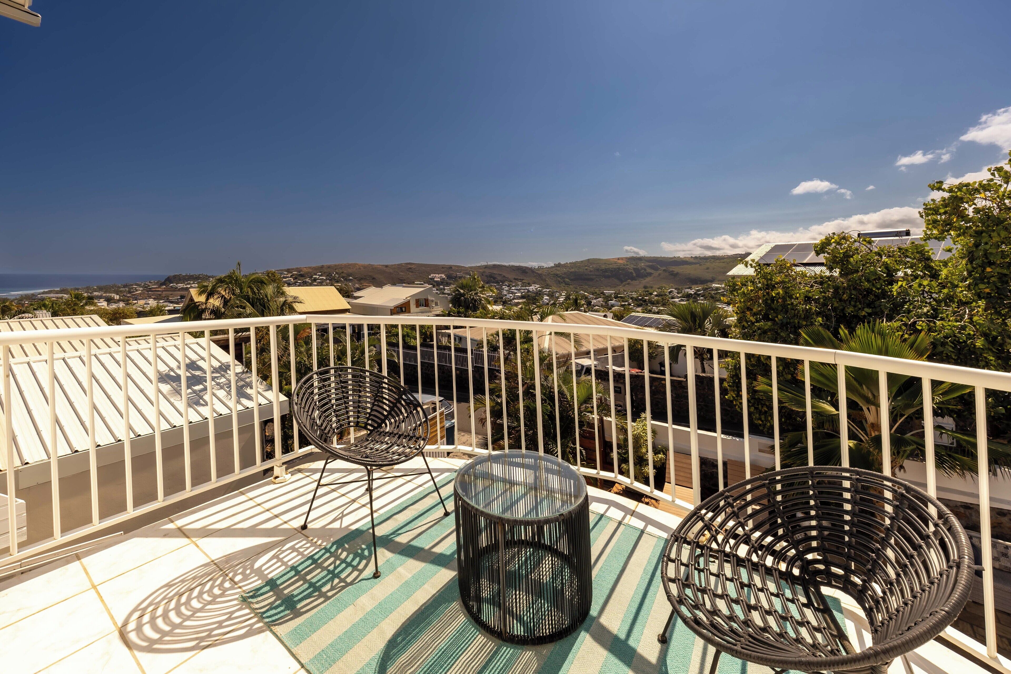 A balcony with a view of the ocean and hills, featuring two wicker chairs and a small table.