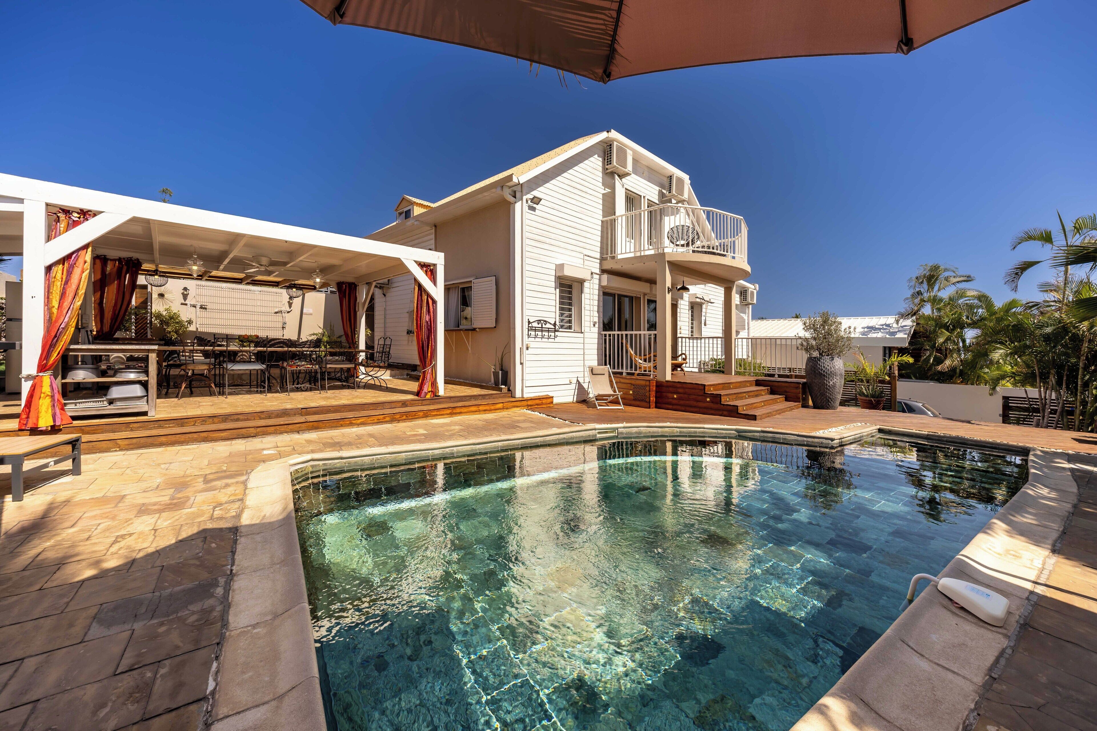 Un espace piscine avec une piscine à eau claire, un patio couvert avec des meubles d’extérieur, et une maison à deux étages dotée d’un balcon.