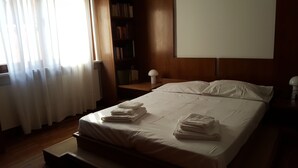 A neatly made bed with folded towels, a bedside table with a lamp, a bookshelf, and a window with curtains. - Large apartment big lounge fireplace two terraces (Venezia)
