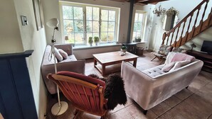 Una sala de estar acogedora con sofá, sillón y una mesa de centro. Amplias ventanas dejan entrar luz natural y ofrecen vistas al verdor exterior.