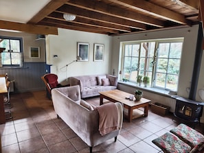 A cozy living room with a sofa, armchair, and a table. There is a large window with a view of trees and a fireplace.