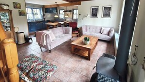 Amplia sala de estar con sofá, una mesa de centro y una mesa de comedor. Se observa una cocina al fondo con armarios azules y una ventana.