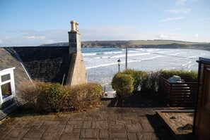 A coastal view from a stone staircase leading to a house with a chimney, overlooking the sea and distant hills.