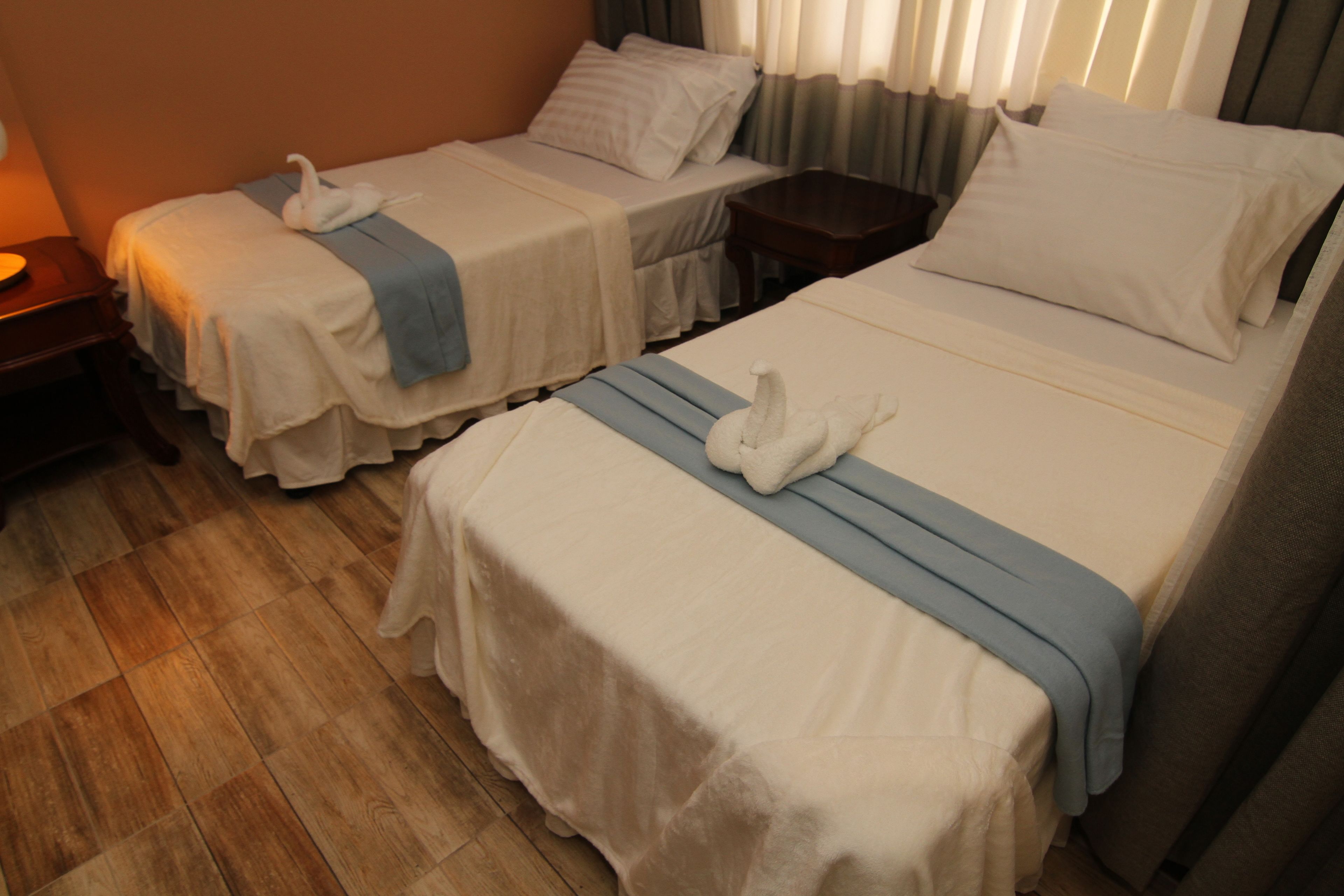 Habitación de hotel con dos camas individuales, cada una con una colcha blanca y un detalle azul. Hay una mesita de noche de madera entre las camas y una pequeña mesa con una lámpara a la izquierda.