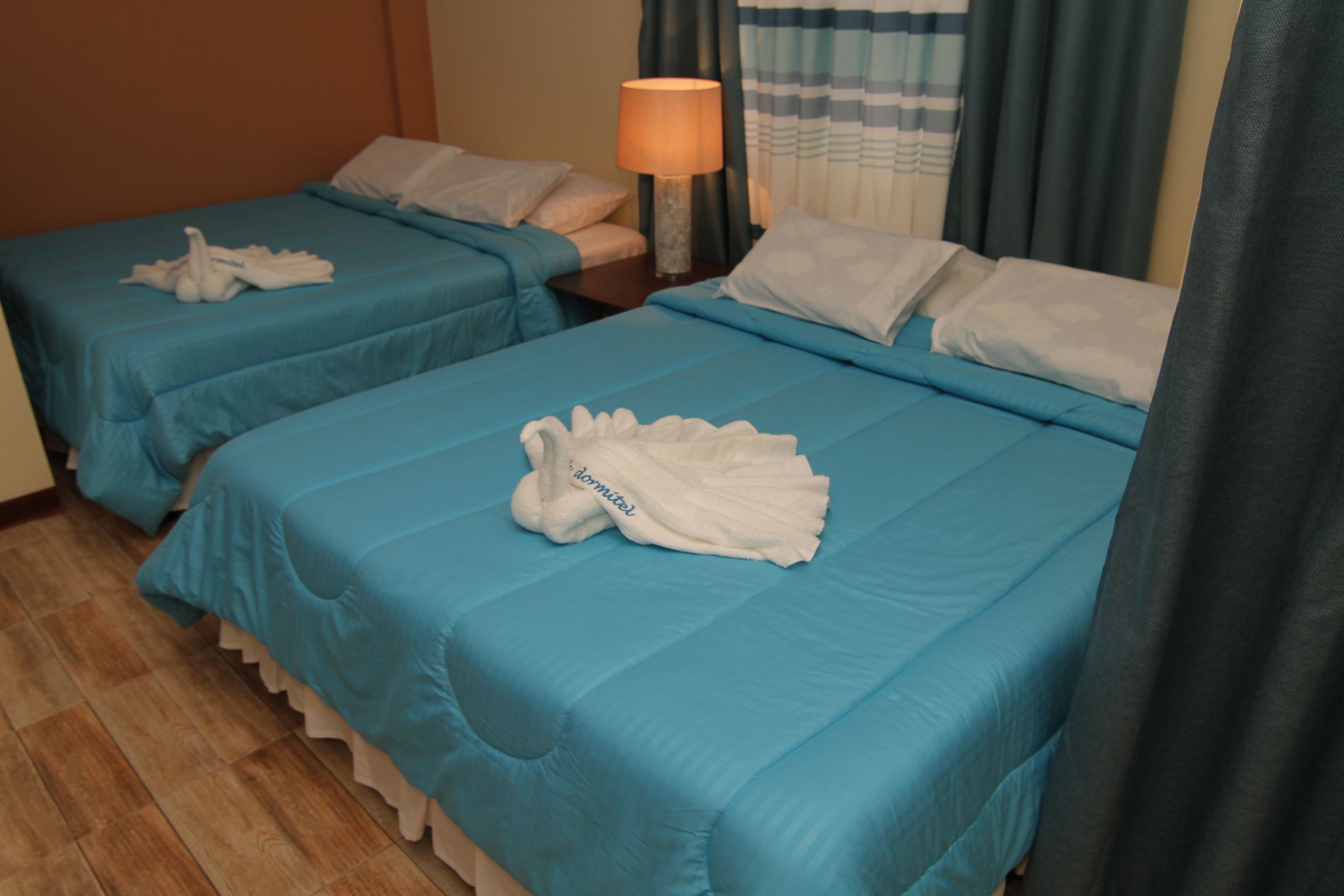 Two twin beds with blue bedding, a bedside table with a lamp, and a towel on each bed.
