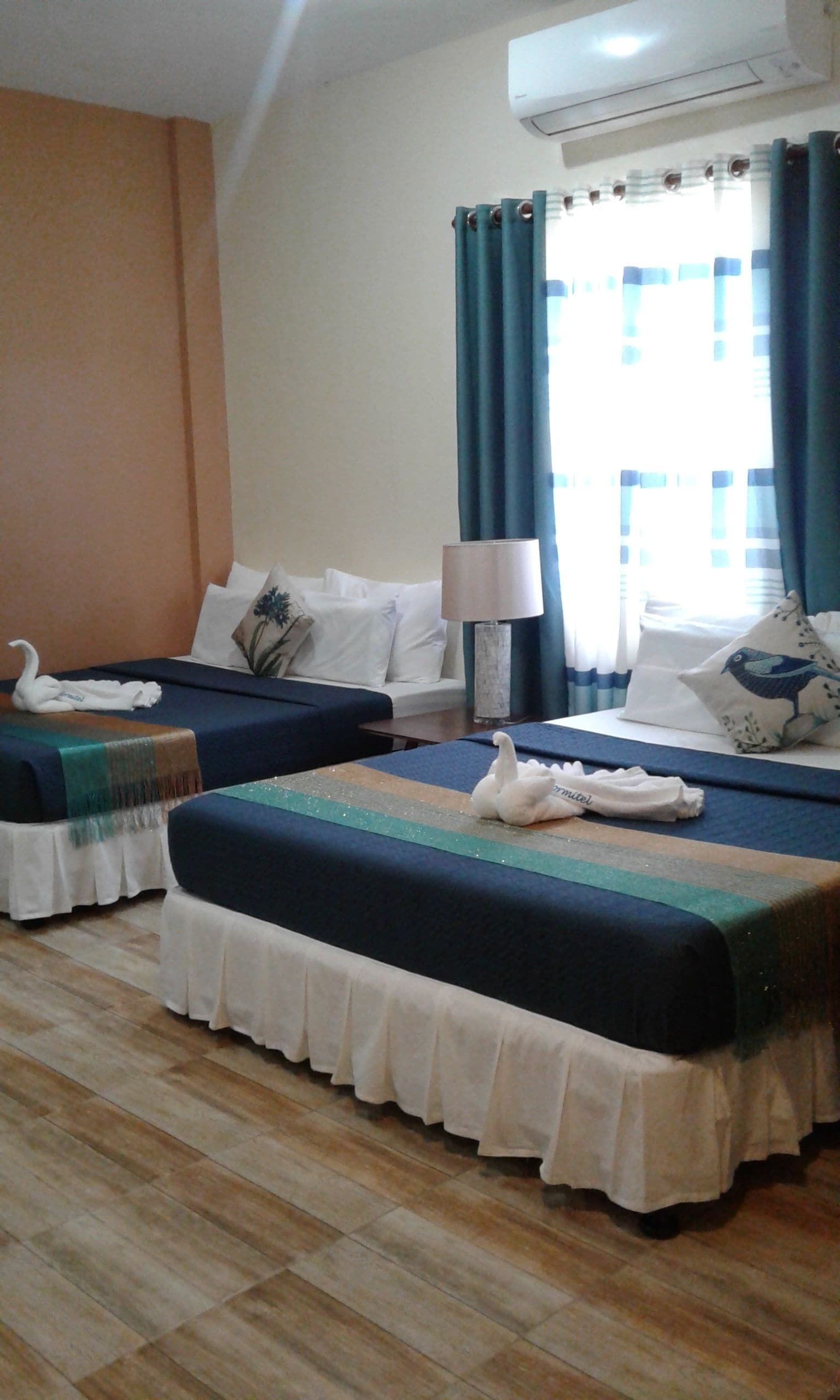 A hotel room with two beds, a lamp, a window with curtains, and an air conditioning unit.