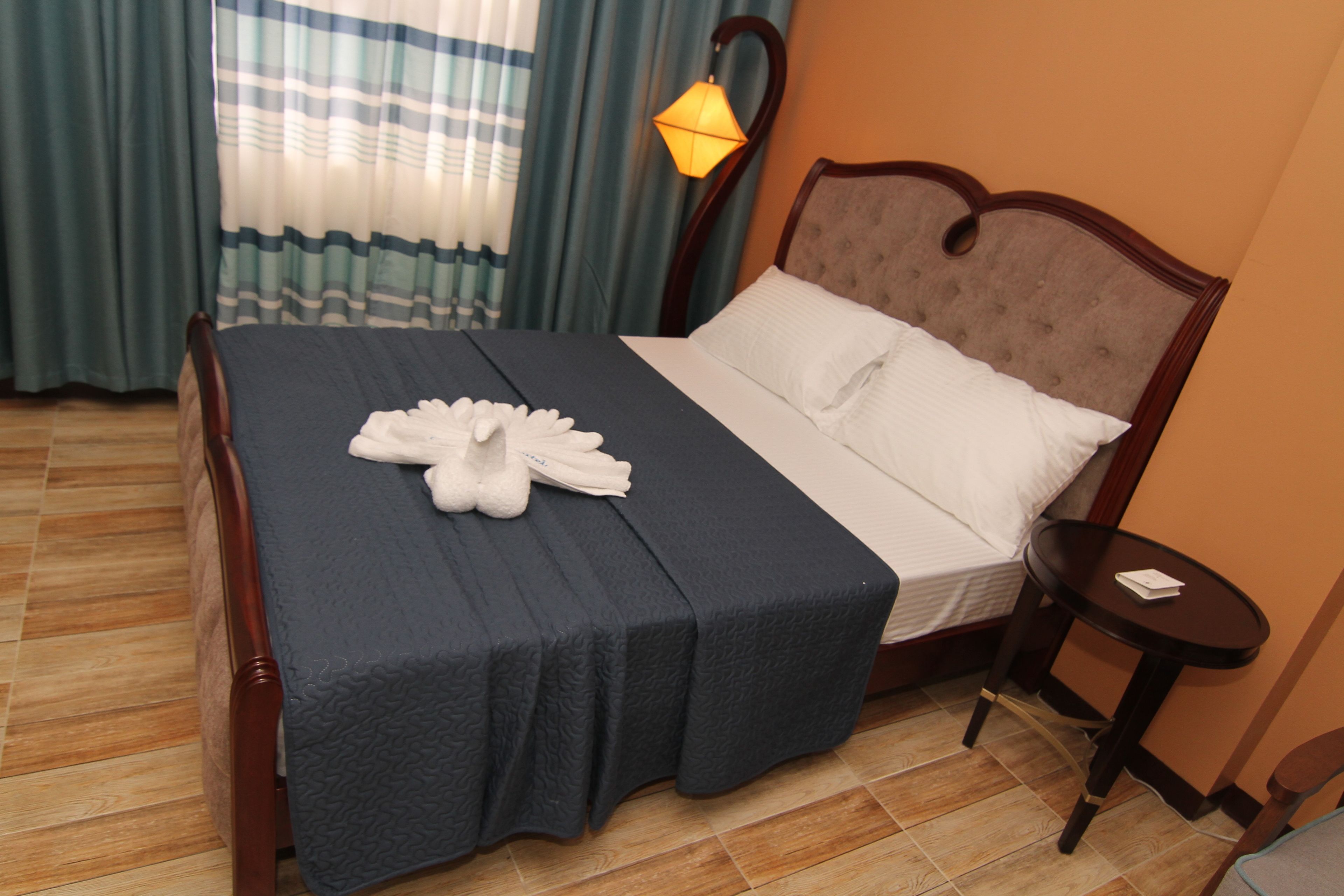 A neatly made bed with a dark blue bedspread and white pillows, a wooden headboard, a small bedside table with a lamp, and a window with curtains.