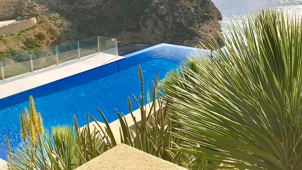 Piscina azul cristalina con barandilla de vidrio, rodeada de vegetación y con vistas a un paisaje costero con casas en una ladera.
