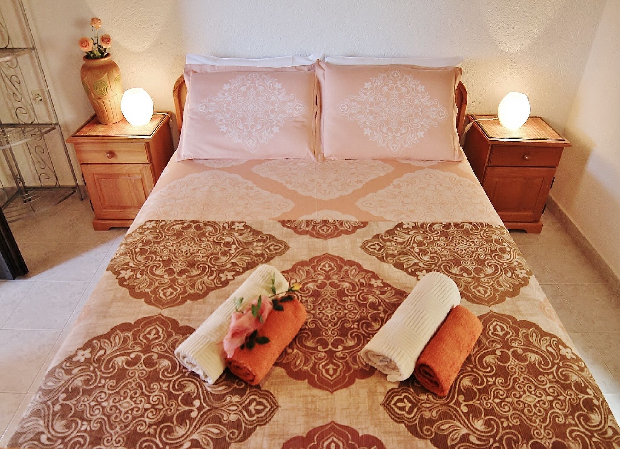 A neatly made bed with patterned bedding, two bedside tables with lamps, and a vase with flowers on one table.