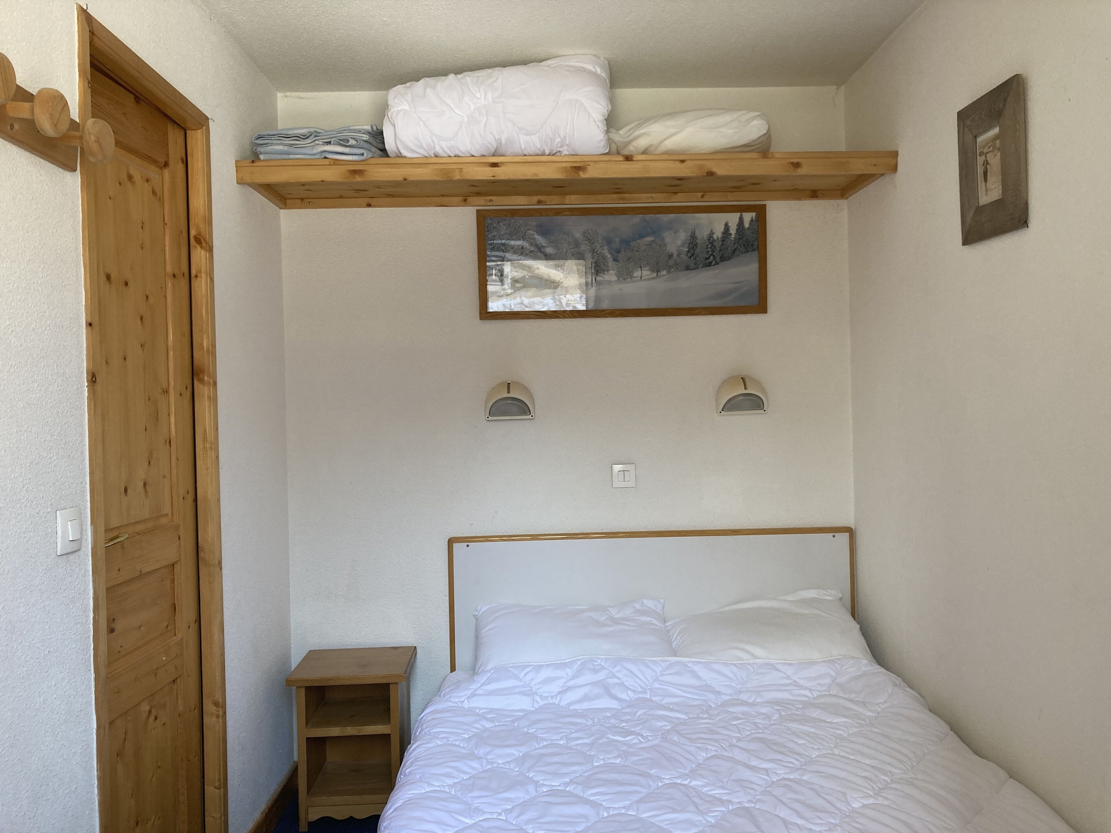 A bedroom with a bed, a wooden shelf with folded towels, a nightstand, and a framed picture on the wall.