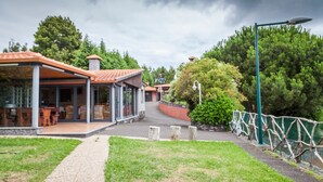 A covered outdoor seating area with tables and chairs, a pathway leading to a building with a tiled roof, and a green fence surrounding the property. - Quinta do Santo by LovelyStay (Santa Cruz)