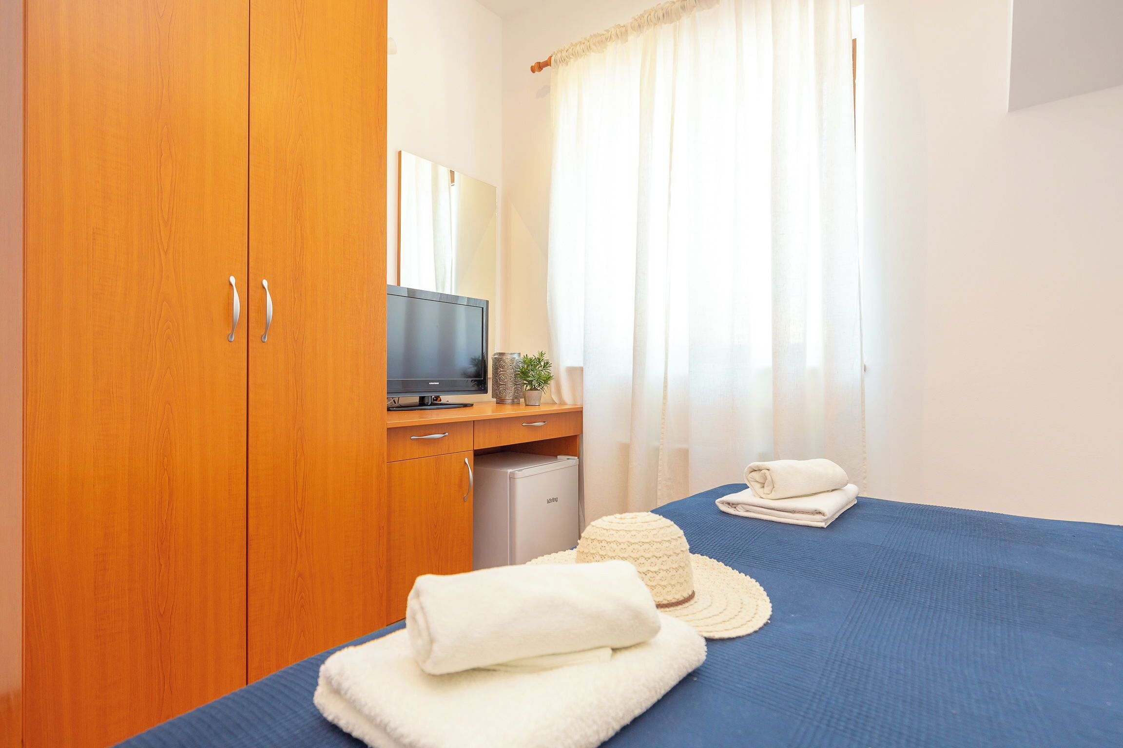 A hotel room with a wooden wardrobe, a bed with a blue bedspread, a television, a small refrigerator, and a window with curtains.