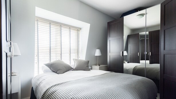 A bedroom with a large bed, a nightstand, a lamp, a window with blinds, and a wardrobe with a mirror.