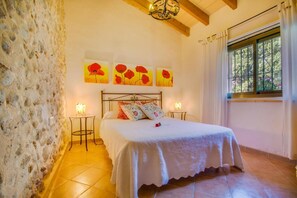 Une chambre avec un mur en pierre, un lit recouvert de linge de lit blanc, deux tables de chevet avec des lampes et trois tableaux représentant des fleurs rouges accrochés au mur.
