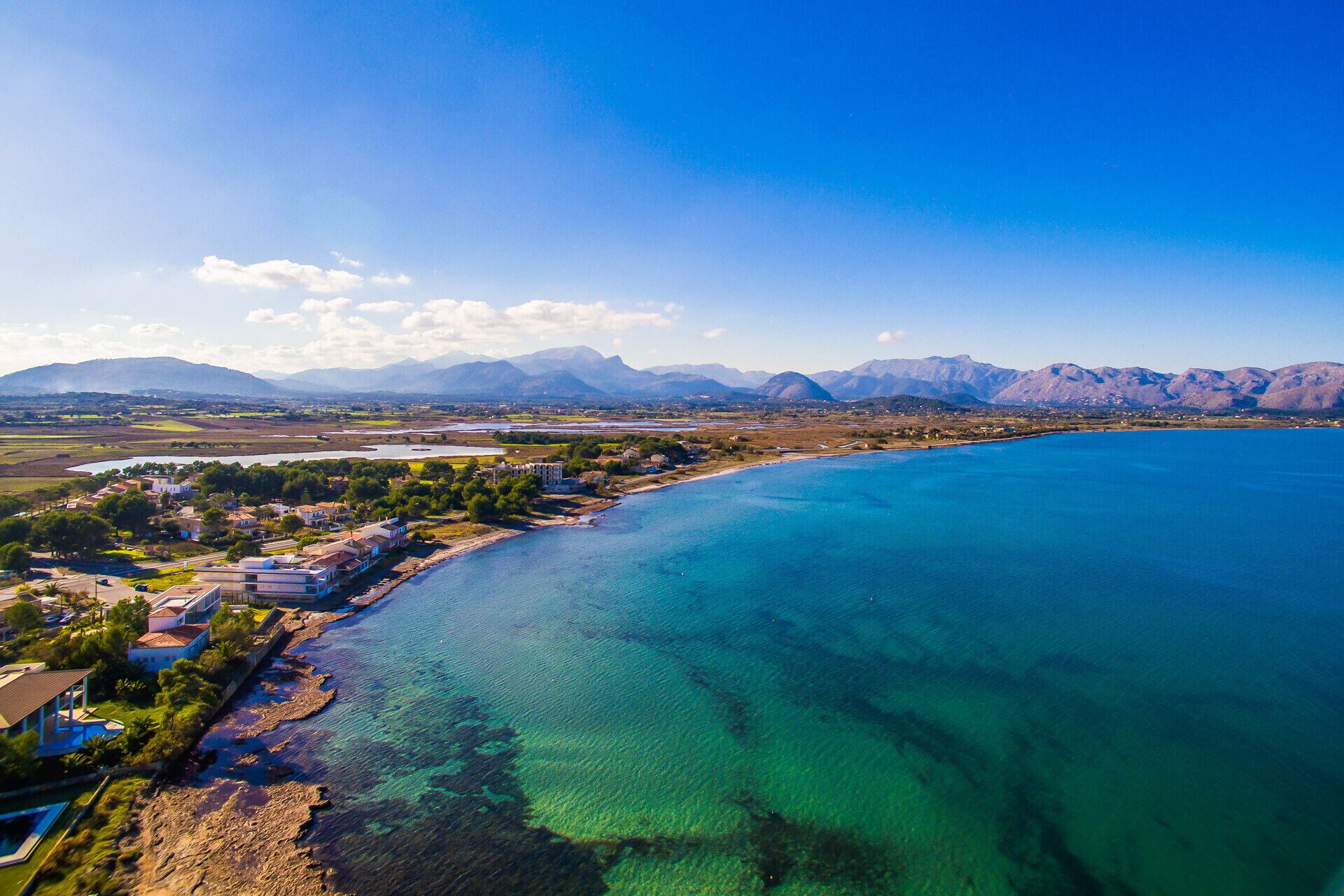 A coastal town with a clear blue sea, mountains in the background, and a mix of residential and commercial buildings.