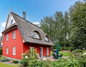 A red house with a thatched roof and a chimney, surrounded by a garden with flowers and a patio area. - Excl. Thatched-roof house with feel-good character in idyllic location, 8 pers. (Glowe)