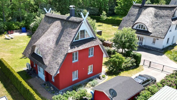 Une maison rouge au toit de chaume, entourée d’un jardin bien entretenu et d’une maison blanche voisine.