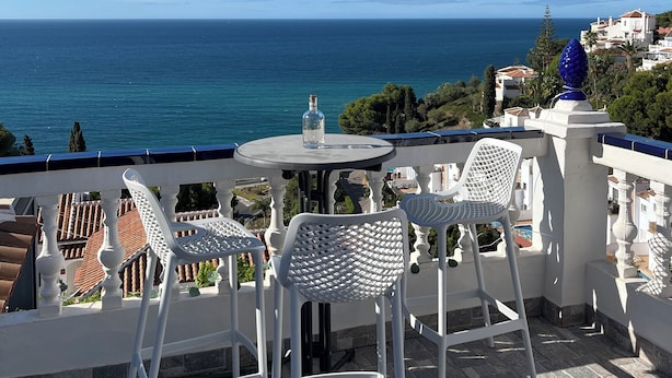 A terrace with white chairs and a table, overlooking a coastal view with houses and the sea.