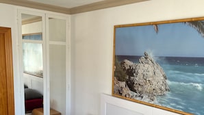 A bedroom with two beds, a large painting of a rocky coastline, a wooden door, and a small bedside table with a lamp.