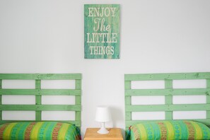 Une chambre avec deux lits en bois verts, une petite table de chevet en bois surmontée d’une lampe blanche, et un mur orné d’une citation inspirante.