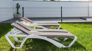 Gartenlounge mit weißen Metallgestellen und dunklen Kissen, auf einer Rasenfläche mit weißer Wand und einem Kaktus im Hintergrund.