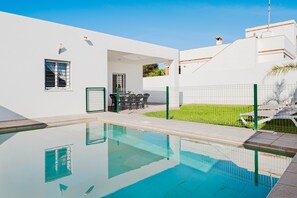 Une piscine à l’eau d’un bleu limpide, un bâtiment blanc doté d’une porte et de fenêtres, et une clôture verte qui entoure l’ensemble.