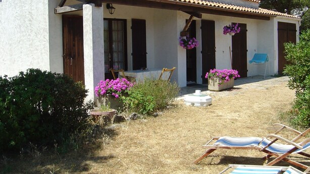 Un bâtiment blanc avec un toit recouvert de tuiles, des portes en bois et des fenêtres. On y trouve deux jardinières ornées de fleurs roses et une chaise bleue à l’extérieur.