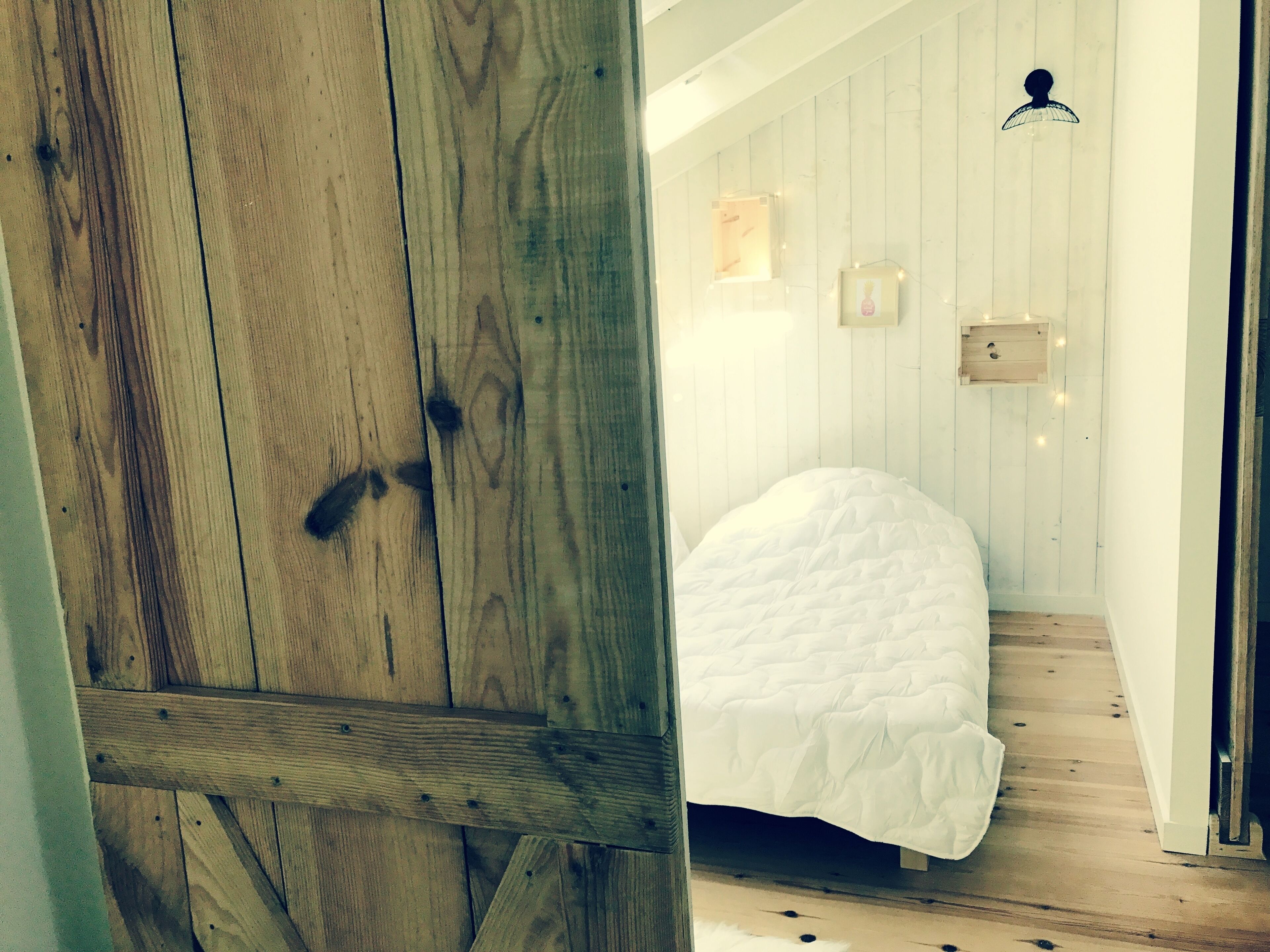 A bedroom with a wooden door, a bed with a white duvet, wooden flooring, and a white wall with a small picture frame.