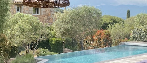 Un espace aménagé au bord de la piscine, avec une terrasse en bois, un bâtiment en pierre en arrière-plan et une vue sur les montagnes.