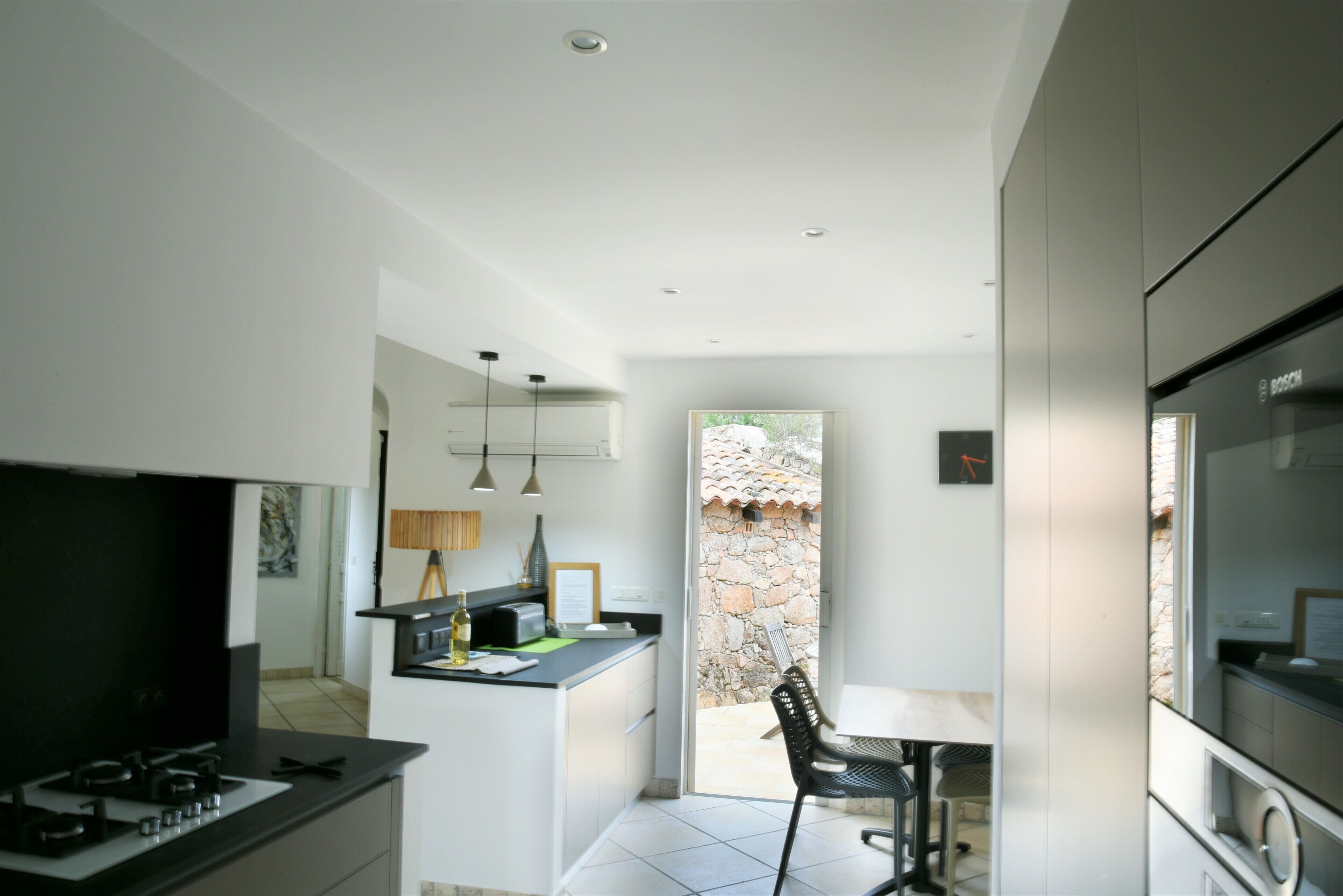 A modern kitchen with a gas stove, a small dining area with a table and chairs, and a view of a stone wall and thatched roof outside.