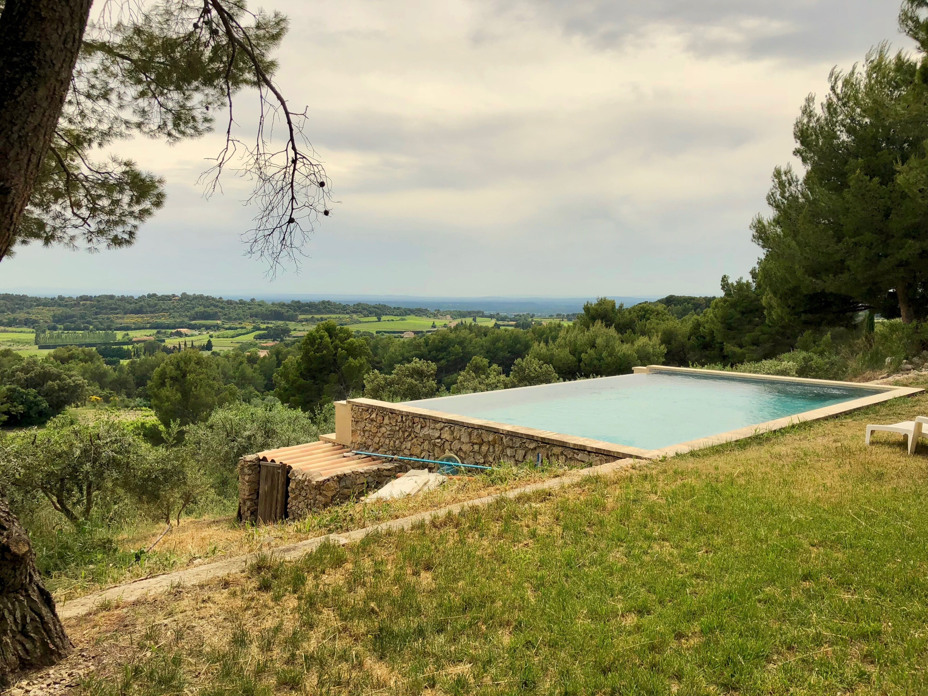 A swimming pool with a stone wall, surrounded by grass and trees, overlooking a scenic landscape.