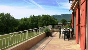 Een balkon met uitzicht op groene bomen en bergen. Er staat een tafel met stoelen en een plant in pot.