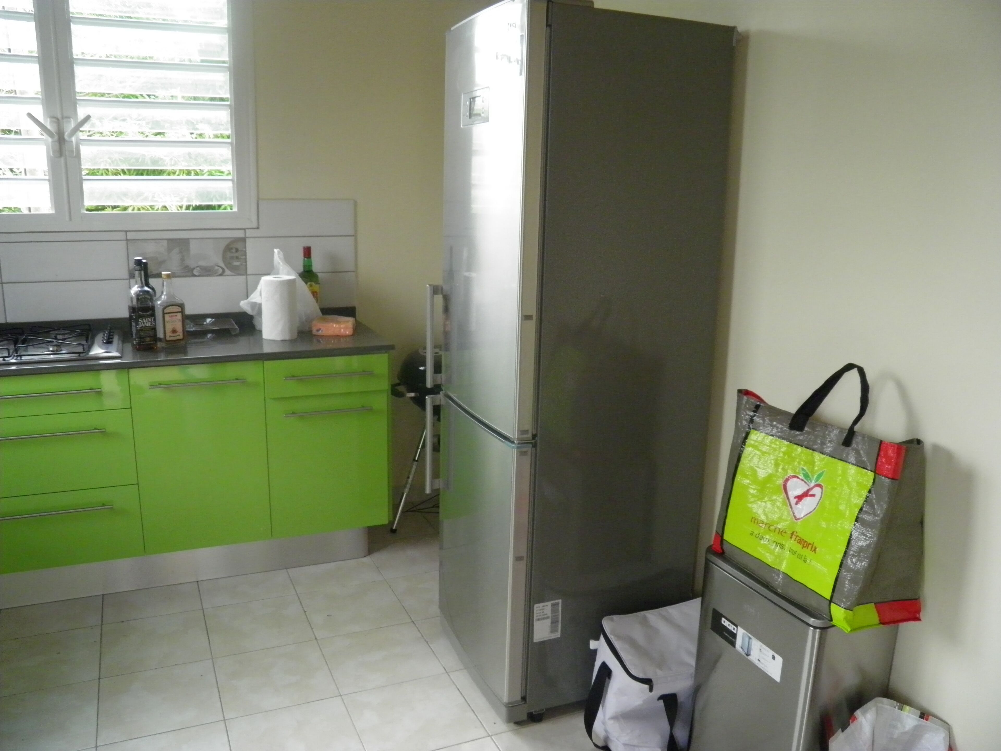 A kitchen with green cabinets, a stainless steel refrigerator, and a window with blinds.