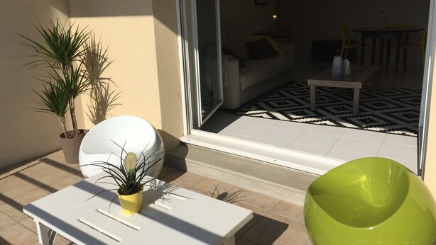 Un balcón con una mesa blanca, dos plantas en macetas y una silla verde.