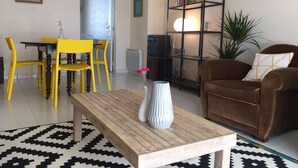 Una sala de estar con una mesa de centro de madera, un sofá marrón, una mesa de comedor amarilla con sillas, una estantería negra y una alfombra estampada.