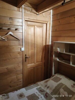 Una habitación con paneles de madera, una puerta, una cama con colcha estampada, una estantería con libros y un perchero con ganchos.