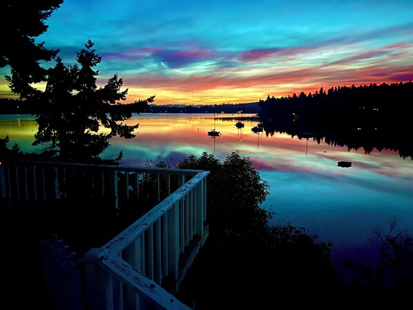A serene lakeside view with a wooden deck overlooking calm waters, boats, and a forested shoreline.