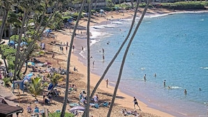 Ein Strand mit badenden und spazierenden Personen, Palmen und Strandsonnenschirmen.