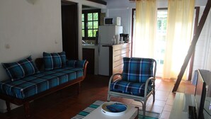 Ein Wohnzimmer mit Sofa und Sessel, einem kleinen Tisch, einer Küchenzeile mit Kühlschrank und Mikrowelle sowie einem Fenster mit Vorhängen.