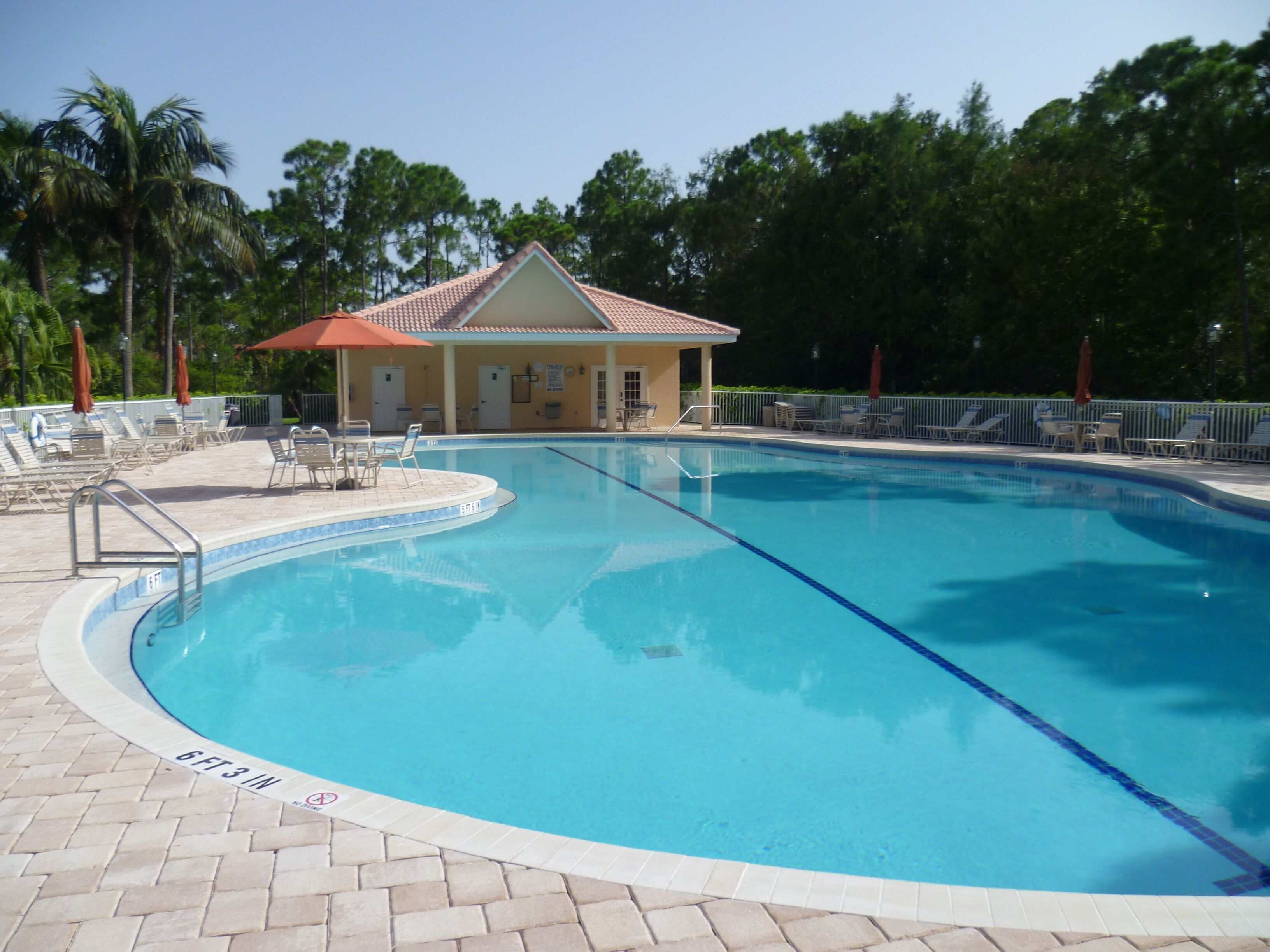 A swimming pool with a tiled deck, lounge chairs, and a building with a red roof in the background.