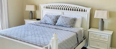 A bedroom with a white bed, two bedside tables with lamps, a ceiling fan, a patterned rug, and a window with curtains.